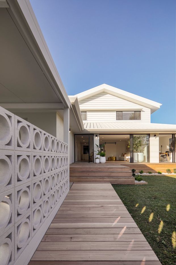 Exterior view of a light-coloured gabled home arranged around a landscaped courtyard. A timber deck and boardwalk lead toward the glazed living area, while a decorative concrete breeze-block screen forms the foreground boundary. The house has horizontal weatherboard cladding, a pale metal roof, and broad indoor–outdoor openings beneath a covered terrace.