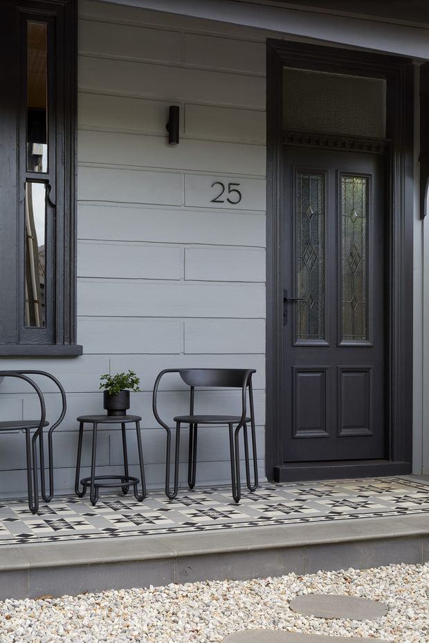 A close view of a refined residential front entry with a dark decorative timber door featuring frosted patterned glass, pale grey horizontal weatherboard cladding, matching dark-trimmed window, house number 25, and a black-and-white geometric tiled porch. Two slim metal chairs and a small plant table sit on the entry landing above a gravel path.