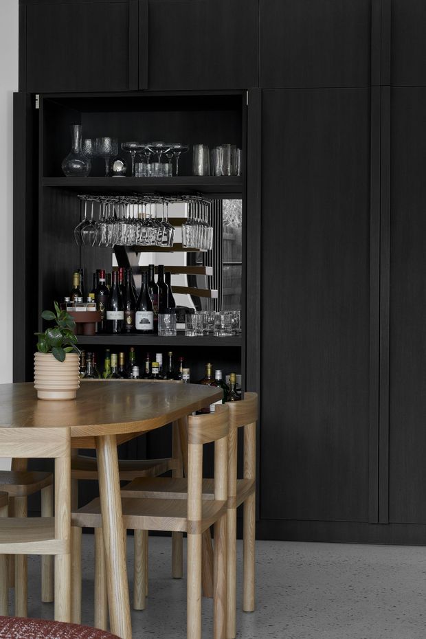 A sophisticated dining and home bar area with a dark timber built-in bar cabinet, open shelving for wine bottles and glassware, and integrated hanging stemware racks. A rounded light timber dining table and matching chairs sit in front, with a small potted plant adding greenery. The space has a minimalist, moody palette of black cabinetry, natural timber furniture, and a grey polished concrete floor.