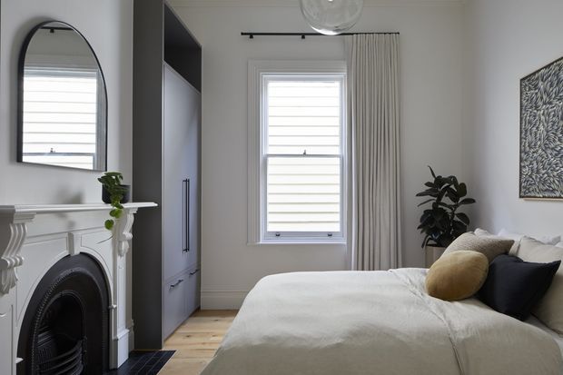 A refined bedroom with a white upholstered bed, layered neutral bedding, dark accent cushions, and a mustard cushion. The room features a traditional white decorative fireplace and mantel, a tall dark built-in wardrobe, pale timber flooring, a large window with white plantation-style blinds and full-length curtains, an arched wall mirror, and a leafy indoor plant. The overall scheme is calm and neutral with a blend of traditional architectural detailing and contemporary furnishings.