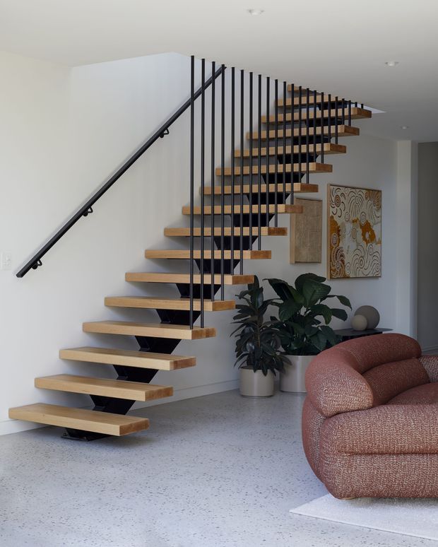 A bright contemporary interior featuring a floating timber staircase with black metal stringers and slender vertical balustrade rods. The stair rises along a white wall beside terrazzo flooring, with indoor plants, abstract wall art, and a curved textured rust-coloured sofa visible in the living area.