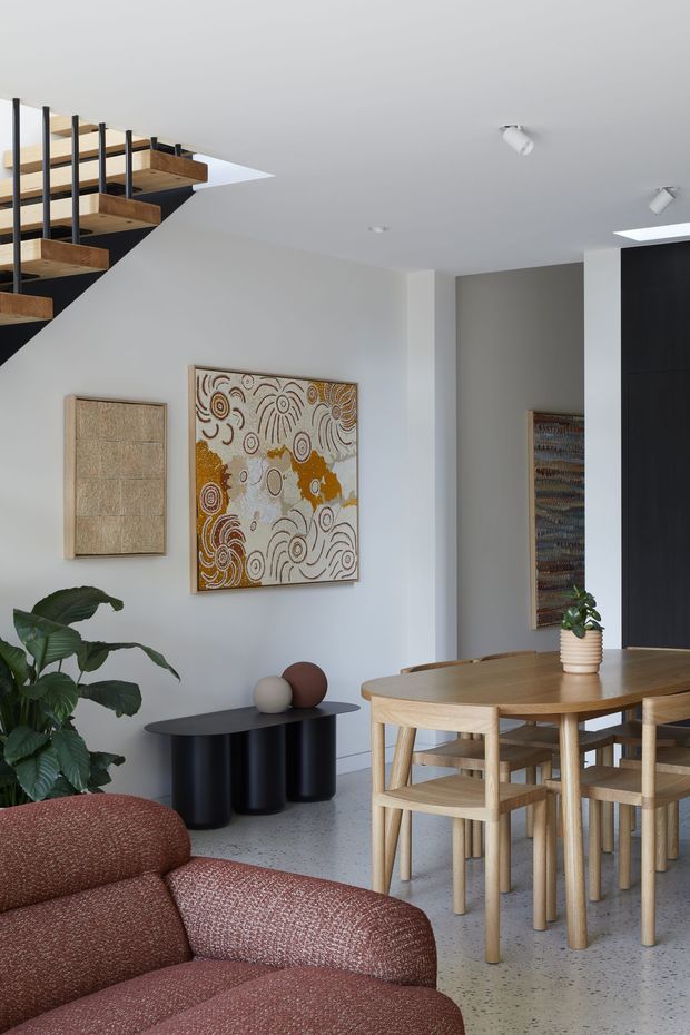 A bright open-plan dining and living area with a timber dining table and chairs, a rust-coloured upholstered sofa, black sculptural console, indoor plant, and Aboriginal-inspired artwork. A striking open staircase with timber treads and slender black vertical balusters occupies the upper left, while concealed cabinetry and recessed spotlights contribute to the clean contemporary interior.