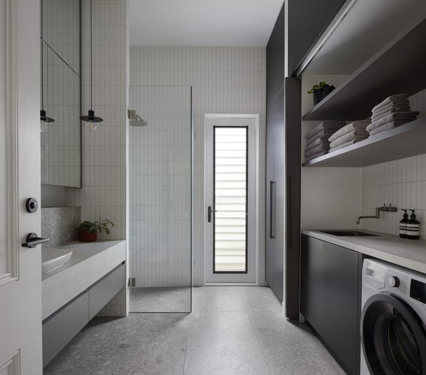 Contemporary compact bathroom-laundry with a walk-in glass shower, wall-mounted vanity, large mirror, vertical white wall tiles, grey stone-look floor tiles, and dark built-in laundry cabinetry. Open shelving displays neatly folded towels, while a front-loading washing machine sits beneath the benchtop. A louvred glass door provides daylight at the rear.