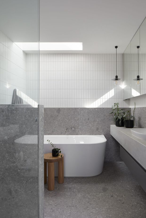 Bright contemporary bathroom featuring a freestanding white bathtub, double vanity, frameless glass shower screen, grey stone-look wall and floor surfaces, and white vertically tiled walls. A skylight and pendant lights provide strong natural and artificial illumination, with small potted plants adding greenery.