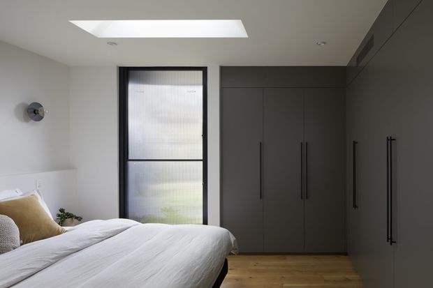 A contemporary bedroom with a low-profile bed dressed in white bedding, a dark upholstered or timber headboard, full-height dark built-in wardrobes, pale timber flooring, and a tall ribbed-glass window. A skylight brings natural light into the otherwise restrained space, complemented by a simple wall light and minimal decor.