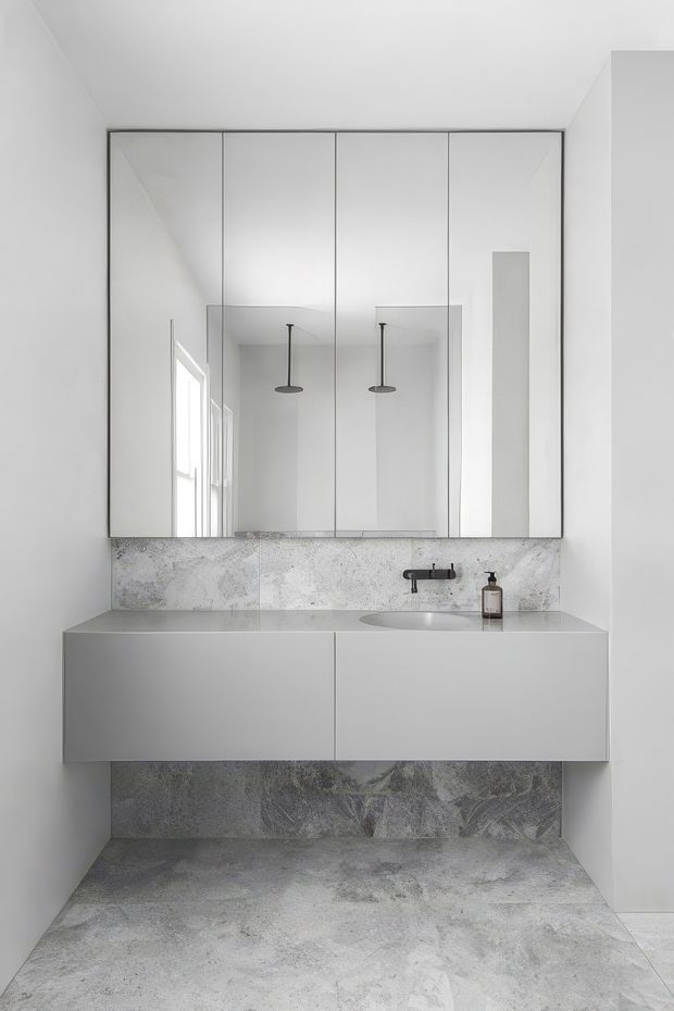 A minimalist contemporary bathroom with a floating white double vanity, integrated basin, wall-mounted dark tapware, large mirrored cabinet fronts, and grey veined stone-look wall and floor finishes. The compact space is bright, clean, and highly refined, with pendant lights reflected in the mirror and a frameless, pared-back fit-out.