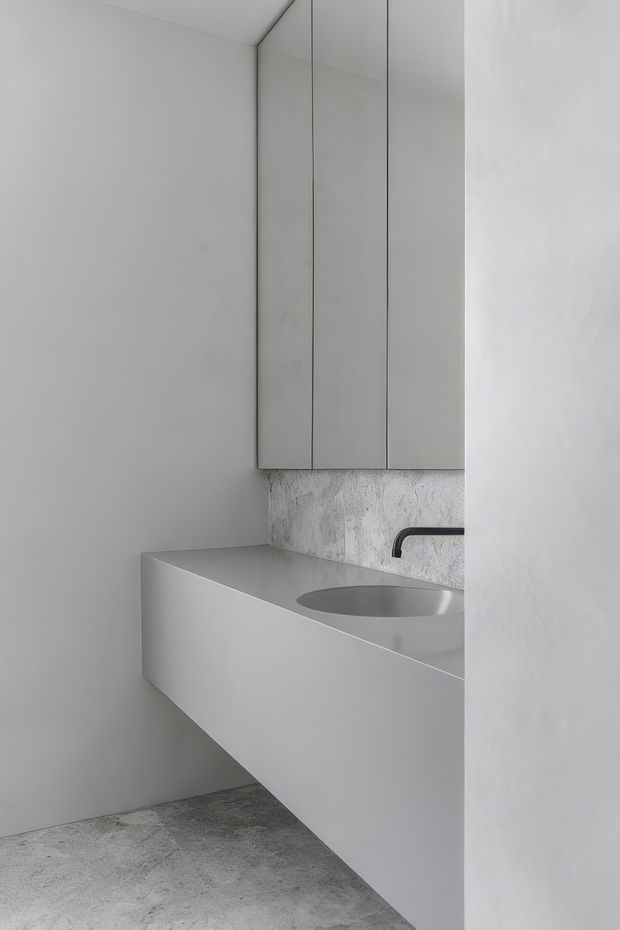 A minimalist monochromatic bathroom vanity with a floating white integrated basin, wall-mounted matte black tap, mirrored or reflective overhead cabinet fronts, and a mottled grey textured splashback. The room uses seamless pale grey-white surfaces and a textured concrete-like floor, creating a restrained contemporary feel.