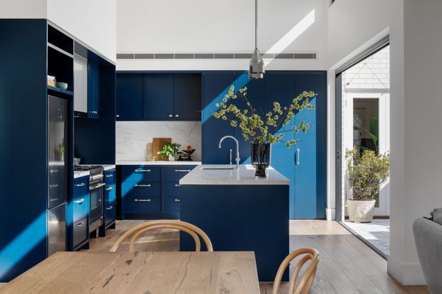 Bright contemporary open-plan kitchen and dining area featuring deep navy-blue cabinetry, a central island with integrated sink, pale stone benchtops, light timber flooring, and a natural timber dining table. Large glazed doors open to a small courtyard, while a linear skylight or clerestory opening and pendant lighting add daylight above the kitchen.