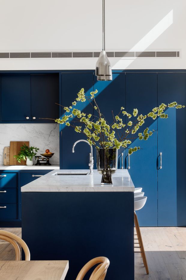 A bright, refined kitchen interior with deep navy-blue floor-to-ceiling cabinetry, a large central island with a white marble benchtop and integrated sink, and a polished metal pendant light. Pale timber flooring and a light stone backsplash provide contrast. A vase of flowering branches sits on the island, with a dining table and curved timber chairs in the foreground.