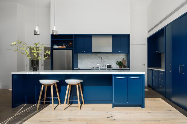 Bright contemporary kitchen with a large deep navy-blue island, integrated seating for two, extensive matching wall cabinetry, stainless-steel appliances, a pale marble-look splashback, light timber flooring, and minimalist pendant lighting. The space appears open, clean, and refined, with a glimpse of adjacent built-in storage.