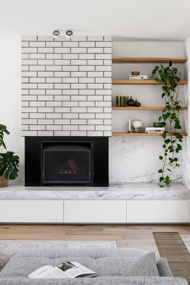 A contemporary living room feature wall with a black fireplace set into a white brick surround, flanked by open timber display shelving and trailing indoor plants. A veined white stone hearth and low white built-in cabinetry run beneath the fireplace, with pale timber flooring, a neutral patterned rug, and a light grey sofa in the foreground.