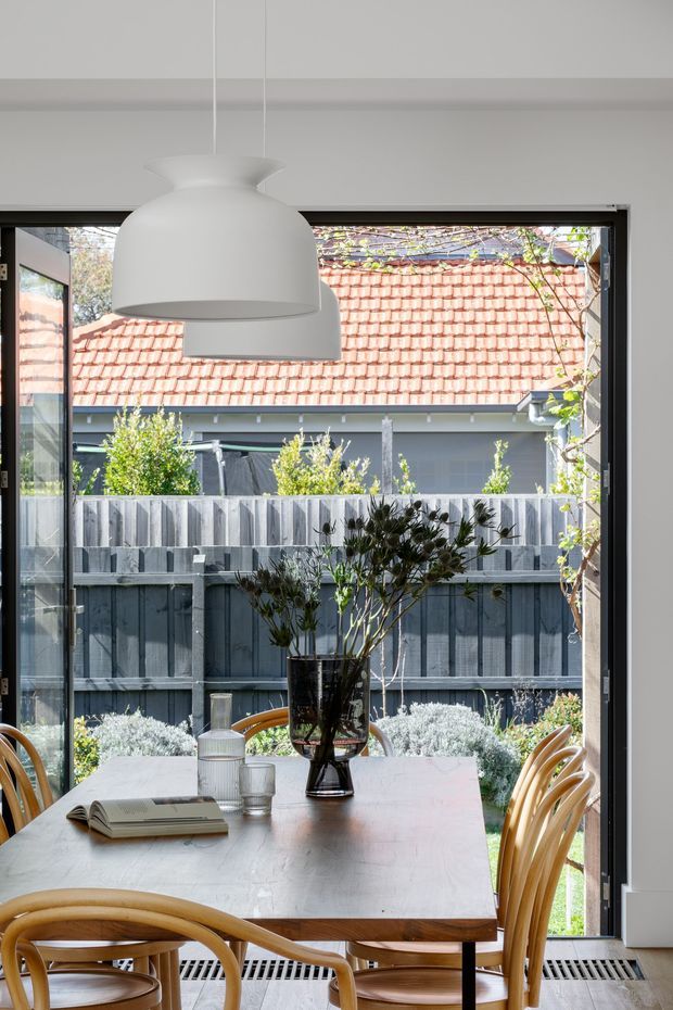 A bright dining area viewed from inside, centred on a timber dining table with curved timber chairs, a dark glass vase with branches, and large white pendant lights. Full-height black-framed glazing opens the room visually to a fenced garden courtyard, with a neighbouring terracotta tiled roof visible beyond. The space has a light, contemporary feel with natural timber accents and abundant daylight.