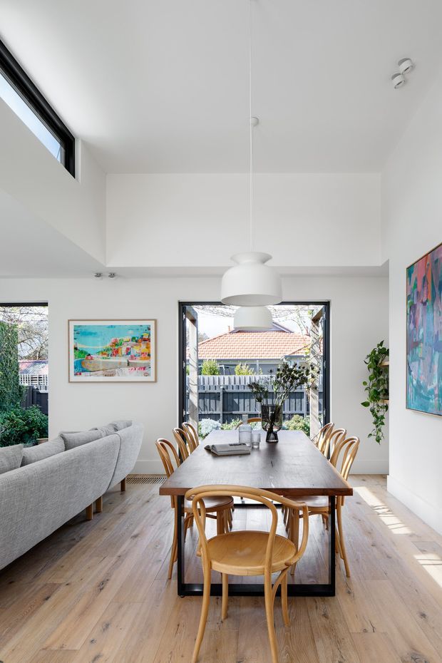 A bright open-plan dining and living area with a large timber dining table, curved timber chairs, grey upholstered sofas, white walls and ceiling, light timber flooring, black-framed windows, pendant lighting, and colourful artwork. The room opens visually to a fenced outdoor area and receives abundant natural light.