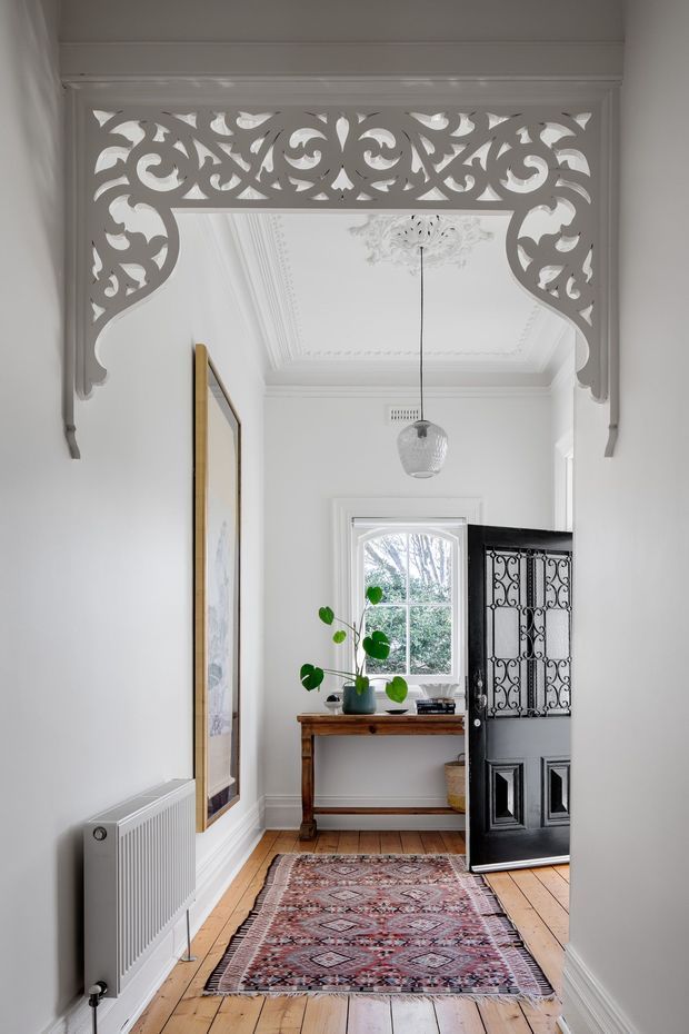 A narrow, elegant residential hallway or entry passage with white walls, decorative ceiling mouldings and cornices, a highly ornate carved white architrave over the opening, pale timber floorboards, and a patterned runner rug. A black glazed-panel door stands open to the right, revealing an entry or adjacent room. A timber console table with a potted plant sits beneath a window, while a framed artwork and white radiator line the left wall. The space combines heritage detailing with restrained contemporary furnishings.