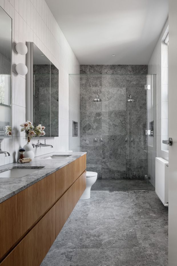 Bright contemporary bathroom with a long timber-look double vanity, grey stone-look benchtop, large-format grey stone tiles across the floor and shower walls, frameless glass shower enclosure, wall-mounted mirrors, and white vertically tiled walls. The room includes twin basins, a toilet, wall-mounted shower fittings, and a white radiator.