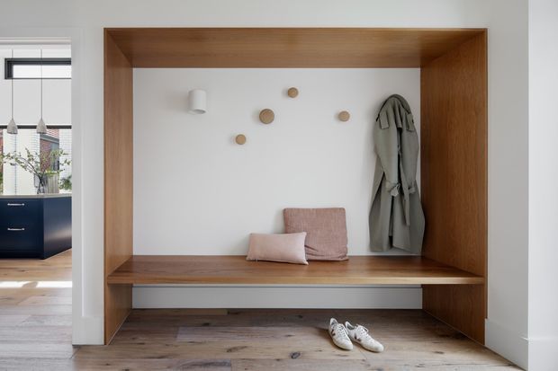A built-in timber-lined entryway nook with a long bench, coat storage, cushions, decorative wall hooks, and shoes on the timber floor. A dark kitchen is visible through the opening to the left.