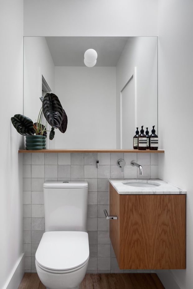 Compact contemporary bathroom with a wall-mounted timber vanity, white basin, chrome tapware, white toilet, square grey wall tiles, and a large frameless mirror. A floating timber shelf with a potted plant and dark soap dispensers adds warmth and contrast. The room has white walls, a wood-look floor, and simple globe lighting.