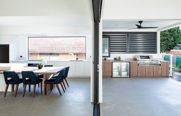 Bright contemporary open-plan living and dining area connected to a covered outdoor kitchen. The interior features a white kitchen with a large window, a timber dining table with dark upholstered chairs, and polished concrete-style flooring. The outdoor zone includes timber cabinetry, stainless-steel barbecue appliances, a bar refrigerator, dark louvred screening, and a ceiling fan.