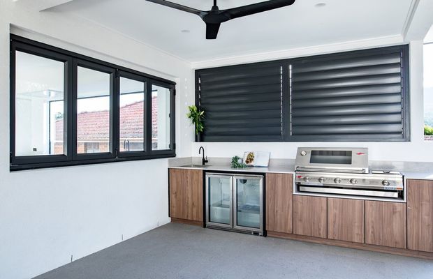 A covered outdoor kitchen with timber-look cabinetry, a stone benchtop, twin under-bench glass-door beverage fridges, a built-in stainless-steel barbecue, sink, dark aluminium-framed glazing, and large dark louvred window shutters. A ceiling fan and tiled or concrete-like flooring complete the functional entertaining area.