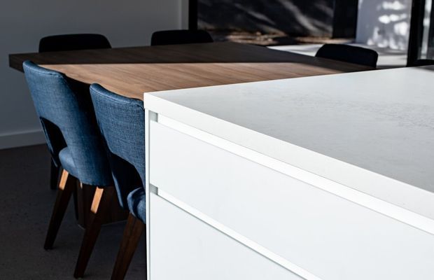 Close-up interior view of a contemporary dining area and adjacent white kitchen cabinetry. A dark timber dining table is surrounded by upholstered navy dining chairs, while a large white stone-look benchtop and handleless drawer fronts dominate the foreground. Dark flooring, neutral walls, and strong natural light create a refined, high-contrast setting.