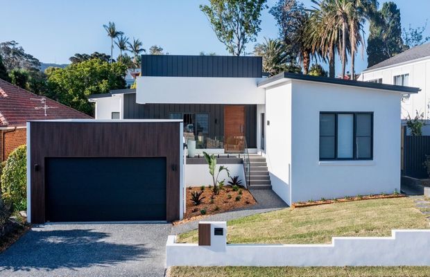 Street-facing photograph of a compact contemporary single-storey home with a white rendered facade, dark metal-clad upper roof volume, timber front door, large dark garage door, and a stepped white boundary wall. The property includes a small landscaped front garden, concrete driveway, and mature tropical and subtropical vegetation in the surrounding suburban setting.