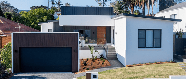 Street-facing view of a compact contemporary single-storey residence with white rendered walls, a flat roof, dark metal cladding to the garage volume, a timber front door, and a stepped entry path. The home is set in a suburban landscaped site with lawn and established planting.