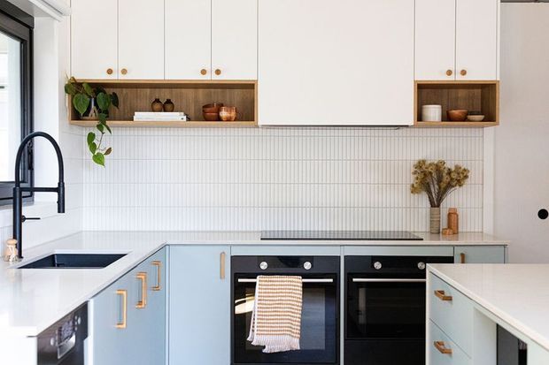 Bright contemporary kitchen with pale blue lower cabinetry, white upper cupboards, warm timber open shelving, a white vertically tiled splashback, dark double ovens, a black gooseneck tap and white benchtops. Natural greenery and simple timber accents add warmth.