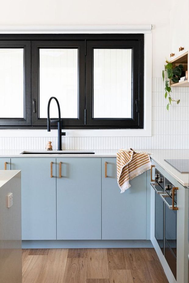 Bright compact contemporary kitchen with pale blue flat-panel cabinetry, slim timber handles, a dark stone or composite benchtop, white vertically fluted wall tile splashback, black-framed windows, and a matte black gooseneck tap. Warm timber flooring and open timber shelving with trailing greenery add contrast.