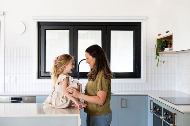 Bright contemporary kitchen with pale blue cabinetry, white tiled walls, light benchtops, and a black-framed window. A woman holds a young child beside the kitchen island. Open shelving with a trailing plant and integrated appliances are visible.
