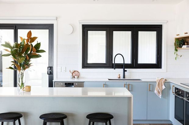 Bright contemporary kitchen with a large pale blue island, white benchtops, black-framed glazing, a black gooseneck tap, integrated appliances and three black bar stools. Vertical white wall panelling and a leafy indoor plant add texture and contrast.