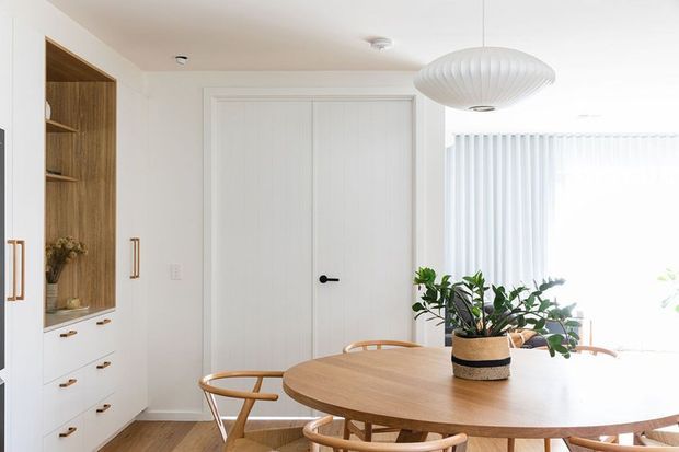 Bright, refined dining area with a large round light-wood dining table, curved timber dining chairs, white walls and cabinetry, a built-in timber-lined display niche, white double doors, vertical blinds, and a sculptural ribbed pendant light. Indoor plants add a natural accent.