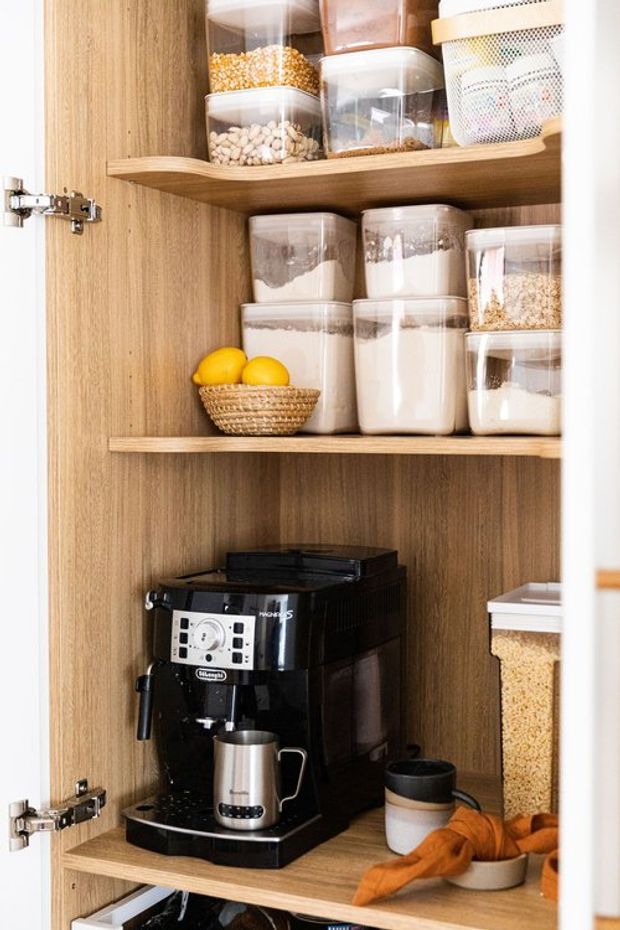 A neatly organised compact pantry cupboard with warm wood-grain shelving and cabinetry. Clear storage containers hold dry goods on multiple shelves, while a black espresso machine, milk frothing jug, mugs and a bowl of lemons occupy the lower shelf. Hinges and white cabinet framing are visible at the sides.