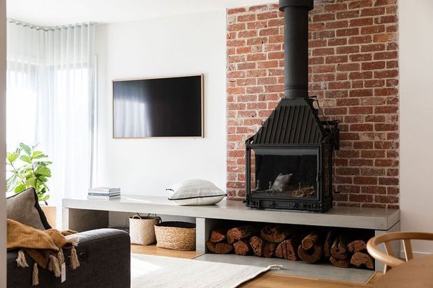 Bright contemporary living room featuring a red exposed-brick fireplace wall, black wood-burning stove with flue, pale stone or concrete mantel, open firewood storage below, wall-mounted television, light timber flooring, and soft neutral furnishings.
