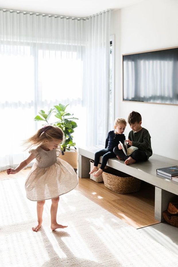 Bright, airy living room with floor-to-ceiling sheer curtains, warm timber flooring, a large pale area rug, and a minimalist built-in bench and media console. Three children occupy the room, with a potted plant and woven storage baskets adding natural accents. A wall-mounted television is reflected in the glossy screen surface.