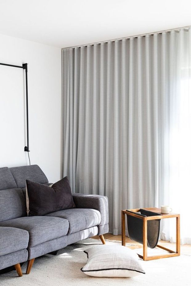 Bright, minimalist living room with a grey upholstered sofa, dark textured cushions, floor-to-ceiling light grey curtains, white walls, a small timber side table, and a large pale floor cushion. Natural light enters from the curtained window, creating a calm contemporary setting.
