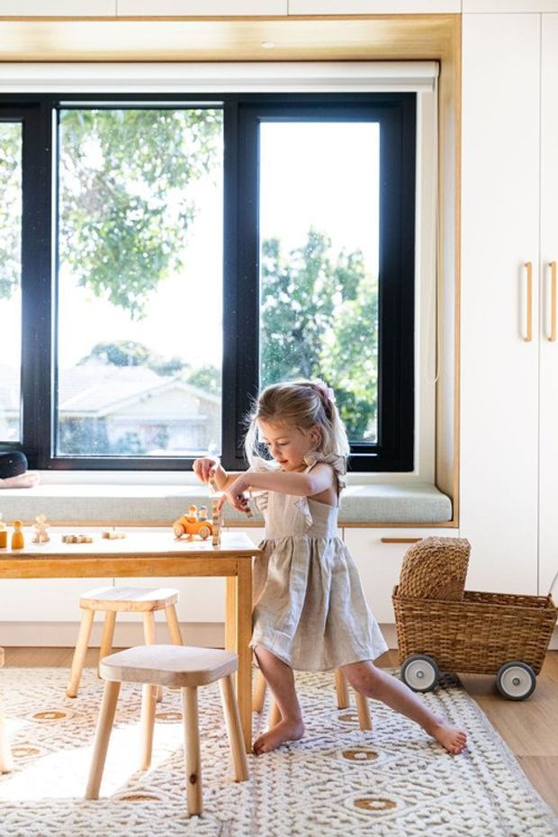 Bright, warmly styled children's play and activity area with a built-in window seat beneath large black-framed windows, white cabinetry with timber trim and handles, a small wooden activity table, stools, woven basket trolley, and patterned rug. A child is playing at the table. The space has abundant natural light and a light timber, white, and neutral palette.