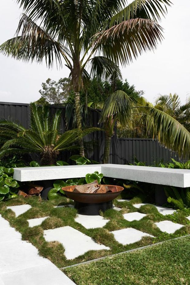 A landscaped outdoor courtyard or garden with tropical palms and lush planting, enclosed by a dark corrugated metal fence. A white angular built-in concrete bench wraps around a rust-toned bowl fire pit, set among irregular white stepping slabs and grass.