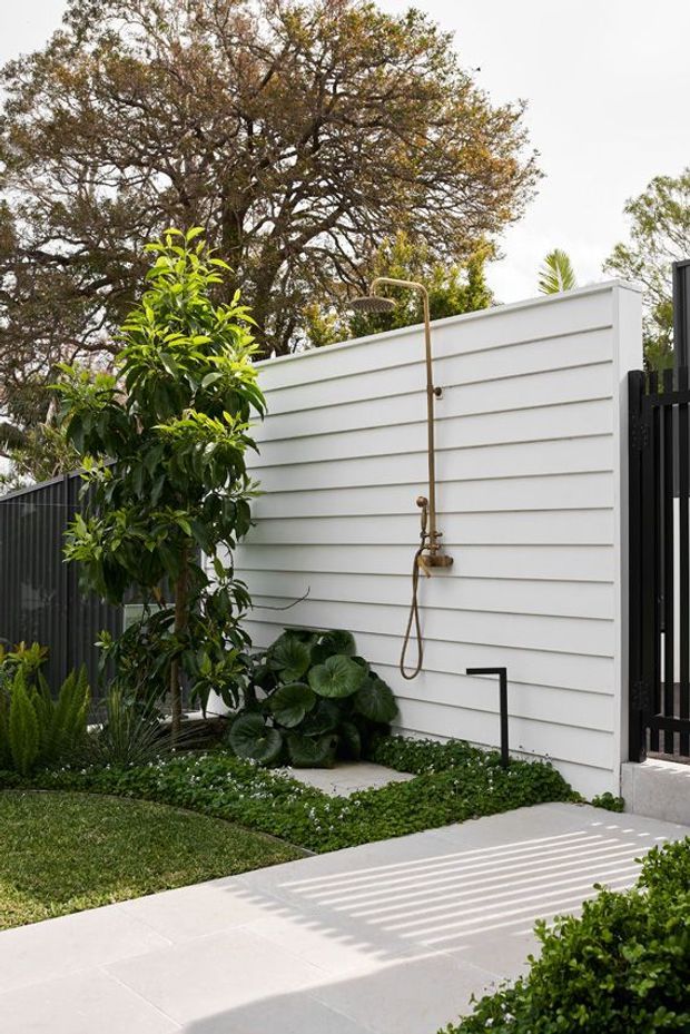 A landscaped outdoor courtyard or garden area featuring an exterior white horizontal-clad wall with a brass-toned outdoor shower, surrounded by lush planting and a pale paved walkway. A black vertical-slat fence and gate frame the right side, with strong striped shadows cast across the paving.