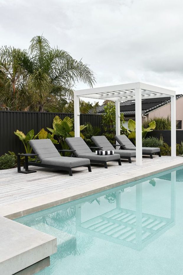 A landscaped residential pool area with a pale timber deck, turquoise swimming pool, and several grey reclining loungers beneath a white rectangular pergola. Tropical planting and a dark privacy fence frame the space, with a tiled-roof house visible behind it under an overcast sky.