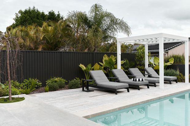 Contemporary outdoor pool area with a pale timber deck, four dark reclining sun loungers, and a white-framed covered pergola. Tropical planting and a dark vertical-panel privacy fence surround the pool, creating a refined resort-style backyard setting.