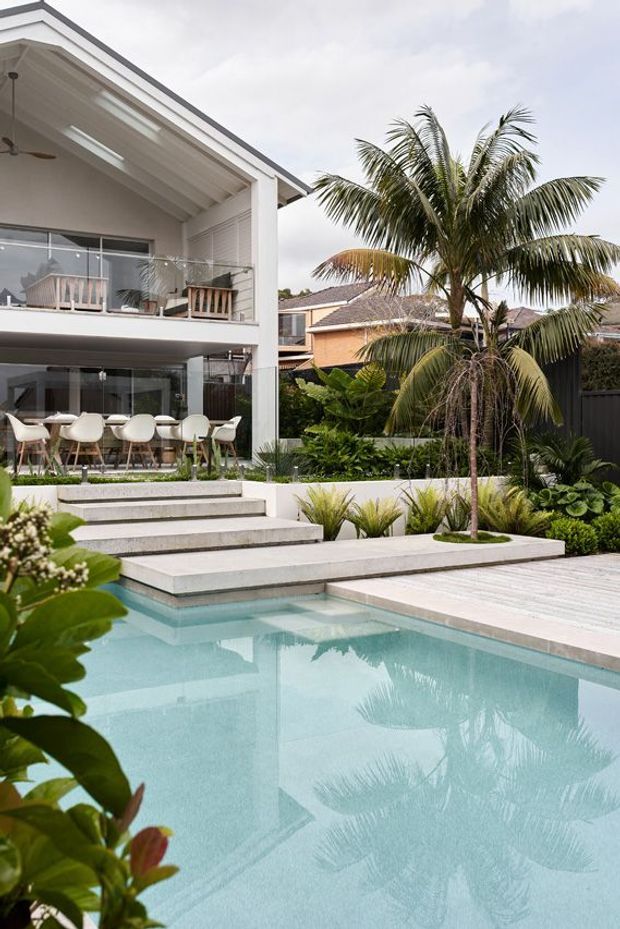 A contemporary two-storey home opens onto a landscaped backyard with a pale turquoise swimming pool, broad concrete steps, and a light timber pool deck. Large glazed openings connect the interior dining area and upper balcony to the outdoor space. Tropical planting, including a prominent palm, creates a lush resort-like setting. The architecture features clean white render, strong horizontal lines, deep roof overhangs, and extensive glazing.