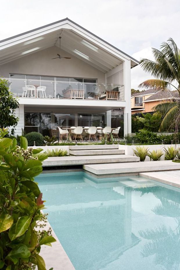 Exterior view of a contemporary two-storey tropical home with a prominent pitched gable roof, white exterior finishes, expansive glass doors and balcony glazing, and a landscaped swimming pool in the foreground. The upper balcony includes outdoor seating, while the lower level opens toward the pool and garden.