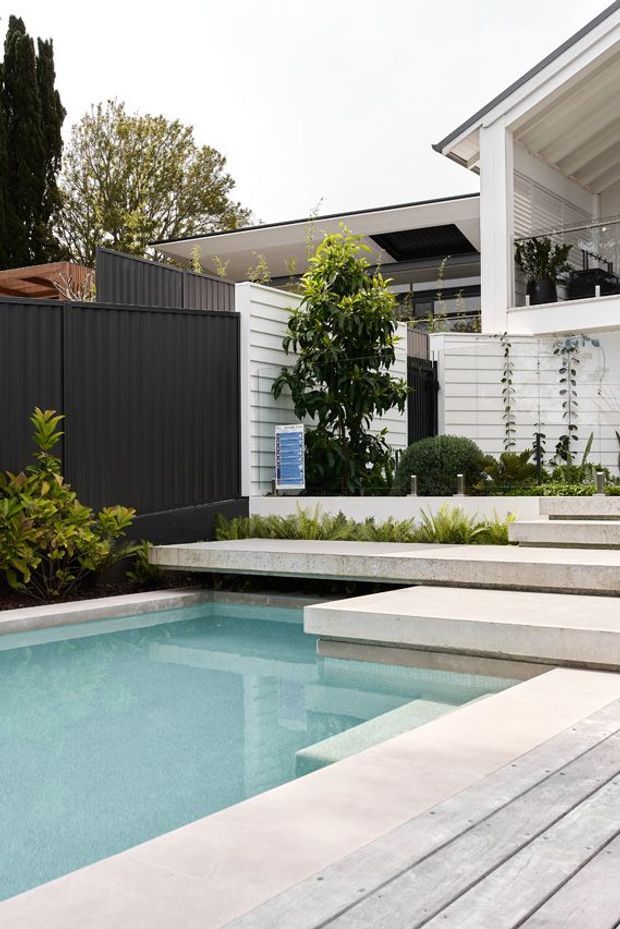 A contemporary landscaped backyard with a geometric swimming pool, pale stone paving, and a light timber deck. Wide floating steps lead from the pool toward a white-clad house and covered outdoor area. Dark vertical fencing, raised garden beds, ornamental grasses, clipped shrubs, and mature planting create a clean, layered courtyard-like setting.