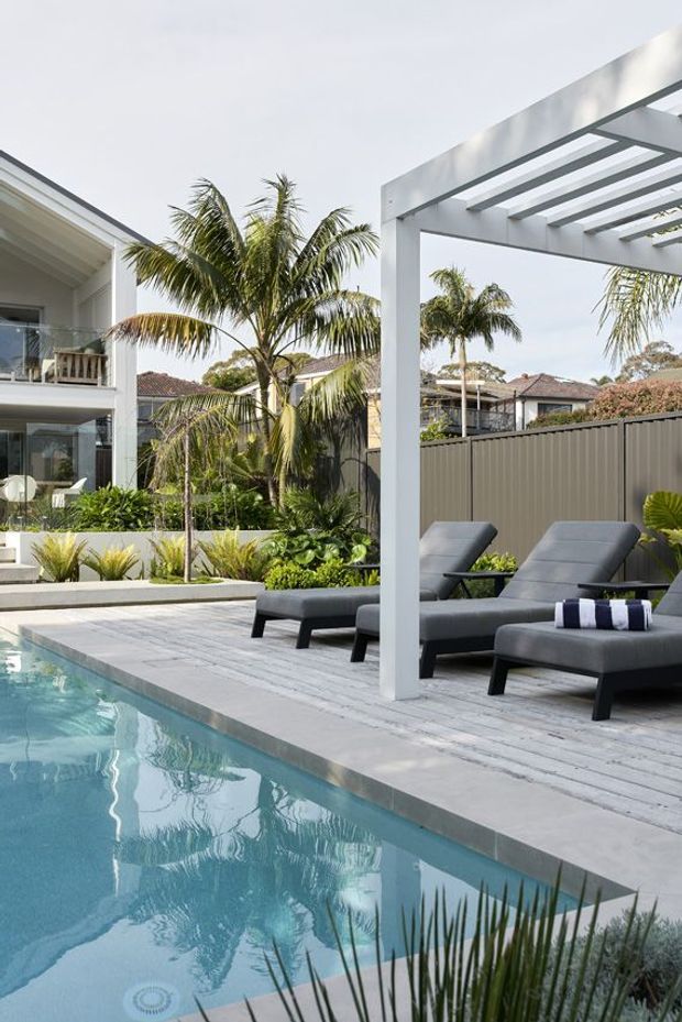 A bright contemporary backyard pool area with a long rectangular swimming pool, pale timber decking, grey cushioned sun loungers, and a white pergola providing shade. Tropical palms, landscaped planting, privacy fencing, and a modern white multi-level house surround the outdoor space.