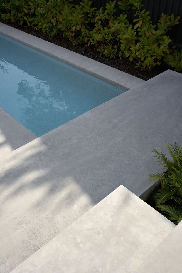 A close-up view of a modern outdoor pool area with pale blue water, large-format grey concrete or stone paving, and light grey pool coping. Dense planted garden beds and dark screening provide a green backdrop, while angular paving edges create a minimalist geometric composition.