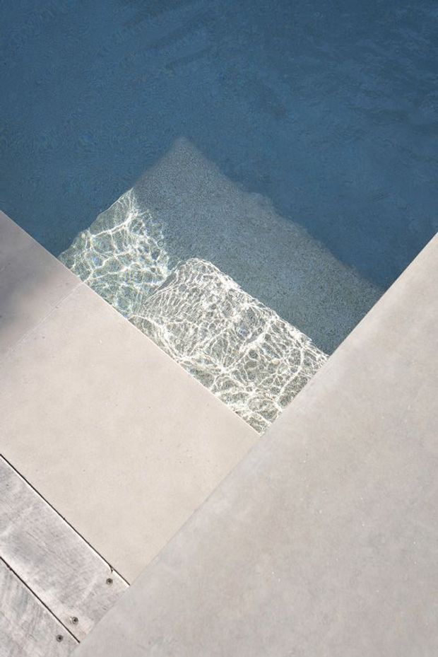 Close-up view of a swimming pool corner with clear blue water, bright rippling reflections, and pale concrete or stone paving around the edge. A narrow timber detail with visible fasteners borders part of the foreground.
