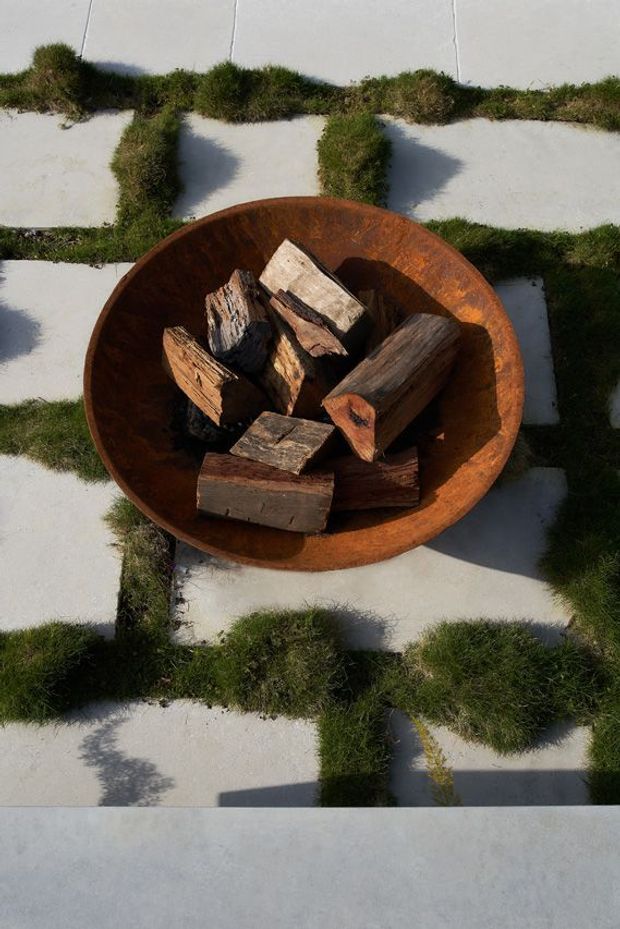 Top-down view of a rusted circular metal fire bowl filled with split firewood, positioned on a grid of pale concrete pavers set into grass. Strong sunlight creates pronounced shadows across the outdoor setting.