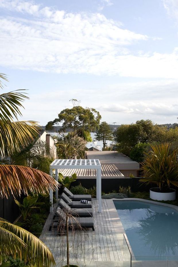 A landscaped outdoor pool area with a narrow rectangular swimming pool, pale timber deck, covered outdoor lounge/pergola, and lush tropical planting. The elevated view looks over rooftops toward a broad body of water, creating a coastal or waterfront setting.