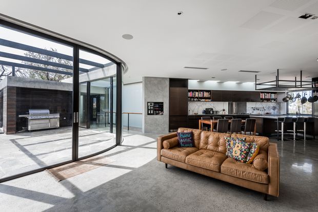Bright open-plan contemporary living area with a large brown tufted leather sofa in the foreground, opening directly onto a paved courtyard through expansive curved black-framed glazing. A modern kitchen with dark cabinetry, island seating, open shelving and pendant lighting occupies the rear of the space. Polished concrete flooring and a minimalist white ceiling reinforce the spacious, refined interior.
