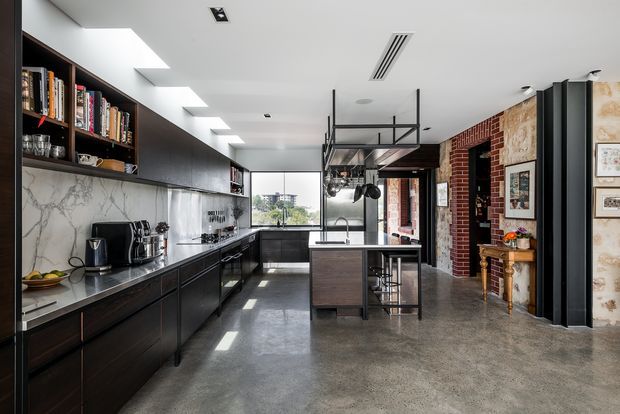 A spacious contemporary-industrial kitchen with dark timber cabinetry running along the left wall, open shelving, a marble-look splashback, polished concrete flooring, and a central island with a sink and bar stools. A large black metal ceiling rack holds hanging cookware above the island. Skylights brighten the room, while exposed red brick, aged masonry, black steel framing, and artwork add an industrial character. A dining or living area is visible beyond the kitchen.